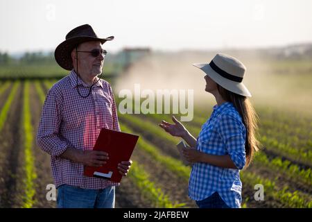 Zwei Bauern, junge Frau und älterer Mann, die im Frühling bei Sonnenuntergang auf dem Sojabohnenfeld sprechen Stockfoto