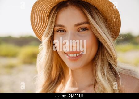 Junge blonde Frau mit Strohhut lächelt und schaut auf die Kamera am Strand Stockfoto