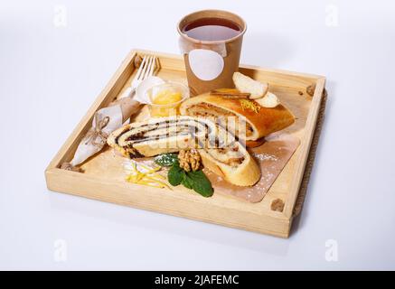 Tee in Papiertasse, Einweglöffel und Gabel, Mohn und Apfelstrudel auf einem rustikalen Holztablett. Weißer Hintergrund, selektiver Fokus. Stockfoto