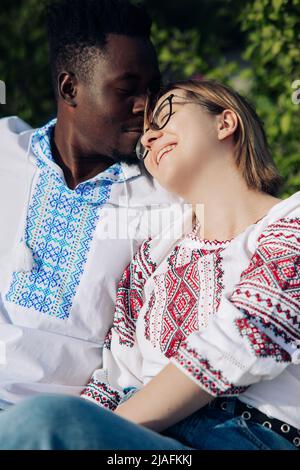 Interrassisches glückliches Paar sitzt auf einer Bank im Garten, gekleidet in traditionell ukrainische ethnische bestickte Hemden. Konzept von Liebesbeziehungen und Einheit b Stockfoto