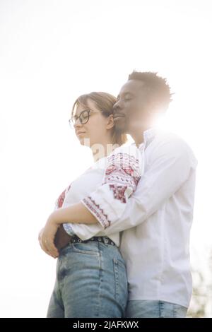 Interracial paar umarmt vor Himmel Hintergrund gekleidet in ukrainische traditionelle ethnische bestickte Hemden. Konzept von Liebesbeziehungen und Einheit Stockfoto