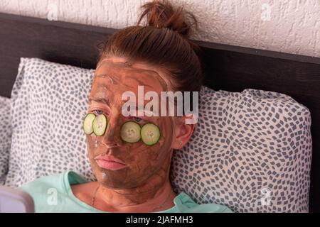 Ein Mädchen in einer kosmetischen Maske und Gurken schneidet sich unter ihre Augen. Eine junge Frau kümmert sich um ihre Gesichtshaut, indem sie Kosmetik anwendet. Stockfoto