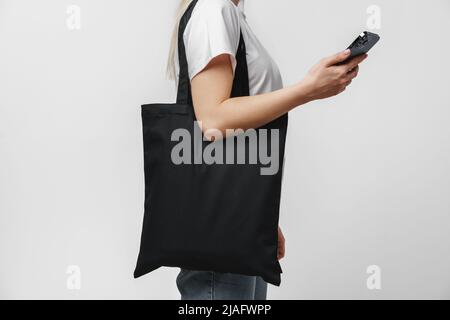 Zugeschnittenes Bild einer Frau mit schwarzer Einkaufstasche aus Baumwolle mit Smartphone vor weißem Hintergrund Stockfoto
