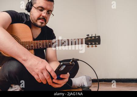 Der junge kaukasische lateinmann mit Brille und Kopfhörer, Musikstudent, sitzt auf dem Boden, seine Gitarre überprüft das Telefon, Kopierraum Stockfoto