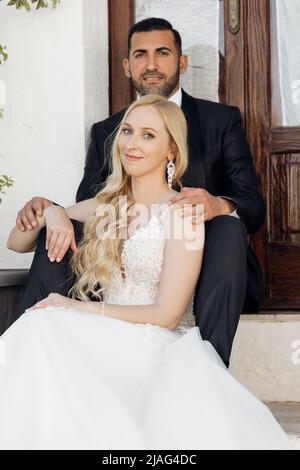 Porträt einer jungen blonden Braut in weißem Kleid und Brunet Bräutigam in Anzug lächelnd und sitzt in der Nähe des alten Stadtgebäudes in Italien, städtischer Hintergrund Stockfoto