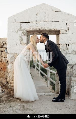 Portrait der blonden Braut in weißem Kleid und Brunet Bräutigam in Anzug küssen und Hände in der Nähe Steinbogen an der Küste in Italien, Seestücke Hintergrund Stockfoto