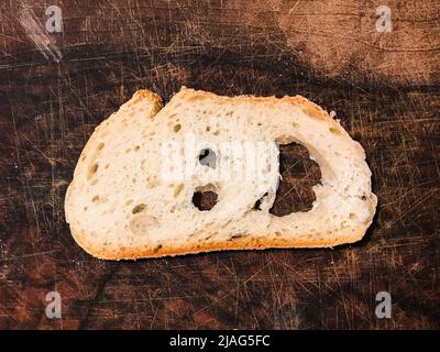 Ein Stück rustikales weißes Brot in Form eines lächelnden Gesichts auf einem hölzernen Schneidebrett Stockfoto