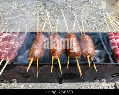 Appetitliche Würstchen und Fleischspieße, die über dem Feuer gegrillt werden, während sie während eines Grills im Freien kochen Stockfoto
