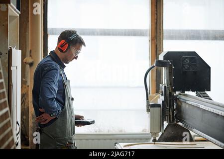 Seitenansicht Porträt eines männlichen Zimmermanns, der die CNC-Schneidemaschine in einer automatisierten Produktionswerkstatt betreibt Stockfoto