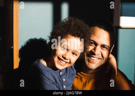 Portrait eines fröhlichen Sohnes mit Arm um den Vater während eines sonnigen Tages zu Hause Stockfoto