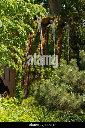 Ein Wasserfall über einer gestreift synthetischen Klippe umgeben von Grün im Medite Smartply Building The Future Garden von Sarah Eberle Stockfoto