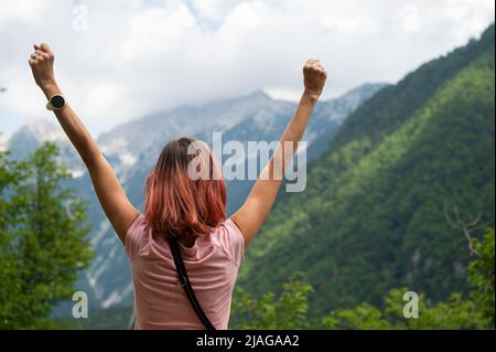 Blick von hinten auf eine junge Frau, Wanderer, die ihre Arme im Triumph nach draußen hebt, und Blick auf die Berge. Stockfoto