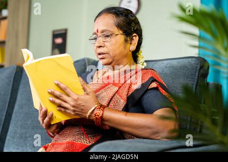 Eine Frau mittleren Alters, die zu Hause mit dem Lesen von Büchern beschäftigt ist, während sie auf dem Sofa sitzt - Konzept von Wissen, Wohlbefinden und Freizeitaktivitäten. Stockfoto