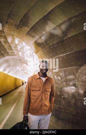 Lächelnder junger Mann, der eine Tasche trägt, während er mit der Hand in der Tasche am U-Bahn-Tunnel steht Stockfoto