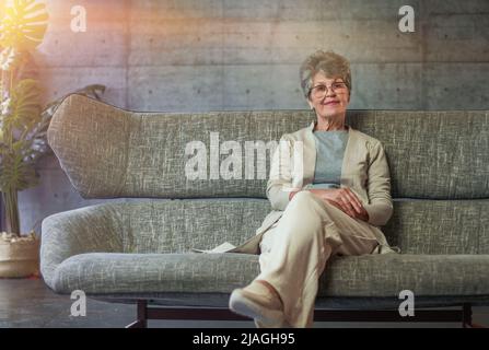 Lächelnde, ältere, grauhaarige Frau im mittleren Alter, die die Kamera anschaut, glückliche alte Dame in einer Brille, die zu Hause drinnen posiert Stockfoto