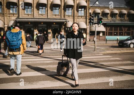 Eine Geschäftsfrau zieht ihr Gepäck, während sie an einem sonnigen Tag die Straße in der Stadt überquert Stockfoto