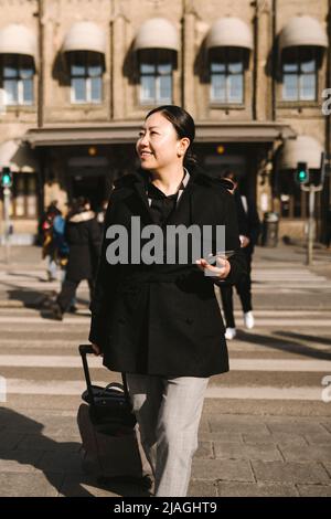 Lächelnde Geschäftsfrau mit Gepäck, das Smartphone hält, während sie die Straße in der Stadt überquert Stockfoto