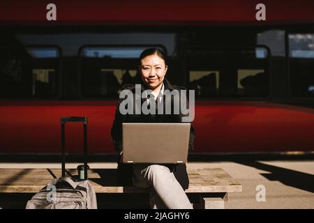 Porträt einer lächelnden Geschäftsfrau mit Laptop auf der Bank vor dem Zug am Bahnhof Stockfoto