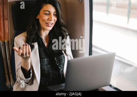 Lächelnde Geschäftsfrau, die Videoanruf auf dem Laptop macht, während sie im Zug am Fenster sitzt Stockfoto