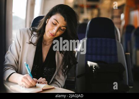 Geschäftsfrau mit langen Haaren, die im Zug auf Tagebuch schreibt Stockfoto