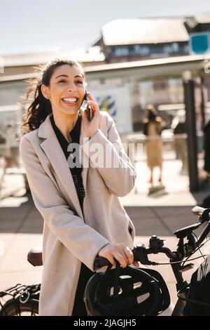Glückliche Geschäftsfrau trägt einen langen Mantel und hält ein Fahrrad, während sie auf dem Smartphone spricht Stockfoto