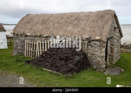 Reetgedeckte Steinhütte mit Torf draußen als Brennstoff für das Feuer und schweren Steinen, um das Dach gegen wildes Wetter zu sichern, auf North Uist Island. Stockfoto