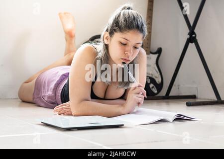 Schöne Latina-Frau, die mit einem Bleistift in der Hand auf dem Boden liegt und auf ihr Notizbuch schaut, das ihre Ernährung berechnet und ausbalanciert. Mädchen neben einem plötzlichen herzstillstand Stockfoto