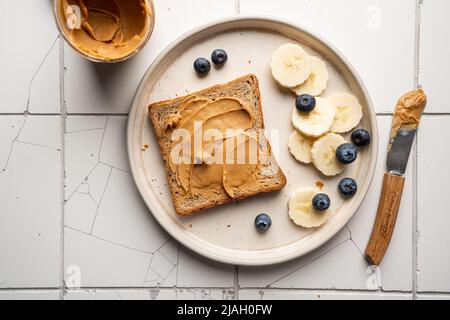 Erdnussbutter Toast mit Bananenscheiben auf Keramikplatte. Gesundes Frühstück oder Snack auf weißem Fliesenhintergrund, Draufsicht Stockfoto