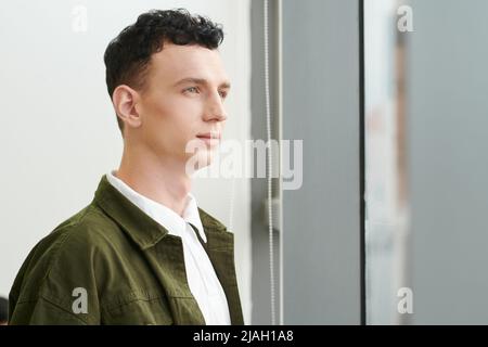 Porträt eines nachdenklichen ehrgeizigen jungen Mannes mit lockigen Haaren, der durch das Fenster nach draußen schaut Stockfoto