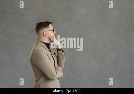 Seitenansicht des jungen Geschäftsmannes, der auf grauem Hintergrund denkt und den Kopierraum ansieht Stockfoto