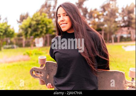 Porträt einer lächelnden jungen Skateboarderin, die ihr Skateboard hält. Frau mit Skateboard im Skatepark und Kamera im Freien Stockfoto