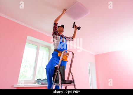 Elektriker wechselt Glühbirne. Junger Mann in Arbeitsuniform, der auf einer Leiter in Wohngebäuden, Büros, Schulen oder Krankenhäusern steht und schraubt Stockfoto