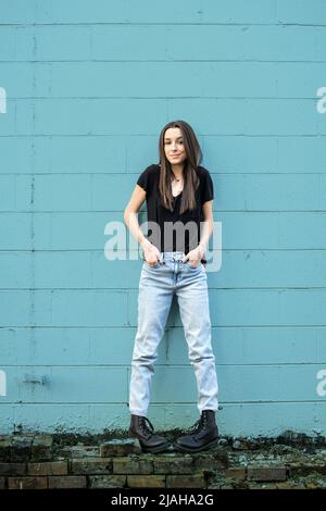 Ein Mädchen im Teenageralter mit langen Haaren, die schwarze, Denim- und Kampfstiefel tragen und sich an eine blaue Blockwand lehnen und mit Attitüde wegschauen Stockfoto