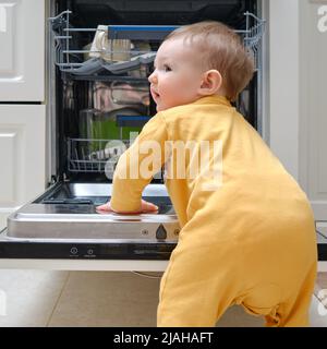 Baby Junge Kleinkind steht bei einer offenen Spülmaschine mit schmutzigem Geschirr im Inneren. Probleme mit der Kindersicherheit im heimischen Zimmer, kleines Kind Stockfoto