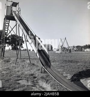 1950s, historisch, draußen auf einem großen Feld, einem Spielplatz und einem kleinen Jungen, der eine lange Metallrutsche hinunterkommt, Margate, Kent, England, Großbritannien. Die Rutsche hat steile Stufen hinauf zu einer überdachten Platte und ein großer Teil der Rutsche hat Seiten. Stockfoto