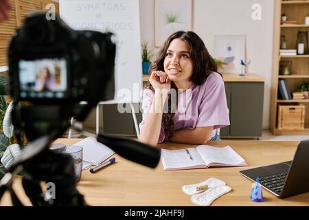 Attraktive junge kaukasische Frau mit langen lockigen Haaren sitzt am Schreibtisch vor der Kamera und spricht über etwas, während sie ein Video für einen Blog aufzeichnet Stockfoto