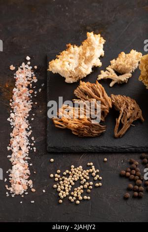 Rohe Sparassis (Blumenkohlpilz), Hericium, Clavaria (Korallenpilz) auf schwarzem Betongrund mit Salz und Gewürzen. Seitenansicht. Stockfoto