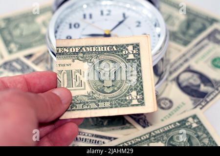 Eine Hand, die amerikanische Geldscheine, USA-Dollar mit einem unscharfen Wecker in Stunden, Minuten und Sekunden im Backgro hält Stockfoto