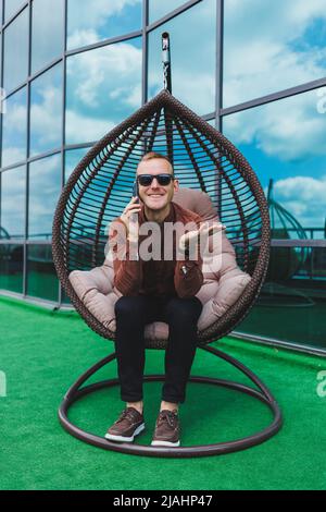 Ein fröhlicher junger Mann in einem Hemd mit Telefon sitzt nach der Arbeit auf der Terrasse eines Bürogebäudes, nutzt WLAN, ein positiver männlicher Mitarbeiter Stockfoto