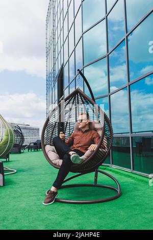 Ein fröhlicher junger Mann in einem Hemd mit Telefon sitzt nach der Arbeit auf der Terrasse eines Bürogebäudes, nutzt WLAN, ein positiver männlicher Mitarbeiter Stockfoto