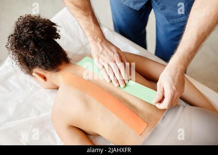 Draufsicht des männlichen Rehabilitationsspezialisten, der Physio-Klebeband auf der Rückseite der jungen Frau aufsetzt, Kopierraum Stockfoto