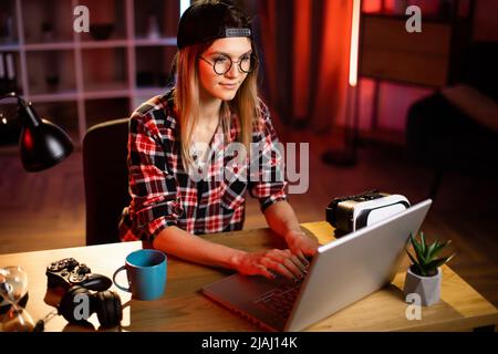 Fröhliche junge Frau in legerer Kleidung, die am Tisch sitzt und auf einem kabellosen Laptop tippt. Junge Frau, die am Abend zu Hause Remote-Arbeit auf einem tragbaren Computer macht. Freiberufliches Konzept. Stockfoto