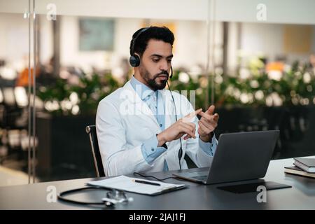 Selbstbewusster, beschäftigter junger muslimischer Therapeut mit Bart in Brille, Kopfhörer und weißem Mantel spricht mit dem Patienten Stockfoto