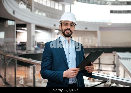 Lächelnd selbstbewusst gut aussehend tausendjährigen arabischen männlichen Ingenieur Geschäftsmann in Schutzhelm, Anzug mit Bart mit Tablet Stockfoto