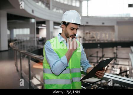 Nachdenklicher junger islamischer Ingenieur-Baumeister berührt seinen Bart in Schutzkleidung und der Hut schaut auf das Tablet Stockfoto