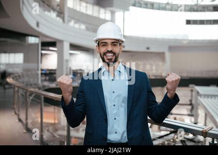 Froh glücklich emotionale junge arabische männliche Ingenieur-Baumeister mit Bart in Hardhat macht Sieg Hand Geste Stockfoto