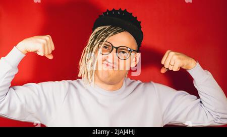 Junger, hübscher Mann mit schwarzer Mütze, Brille mit Händen, die Kraft zeigt. Starker Hipster mit blonden Dreadlocks in Kleidung zeigt Bizeps auf rotem Hintergrund Stockfoto