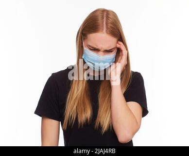 Frau trägt schützende weiße Maske gegen Virus, fühlt starke Schmerzen in Schläfen, hält die Finger an den Kopf. Junges Mädchen mit langen blonde in schwarz t gekleidet Stockfoto