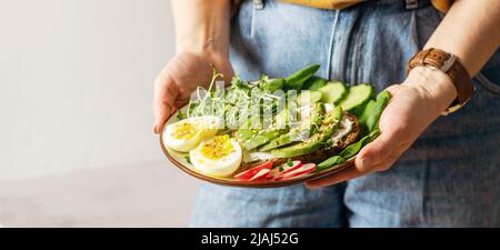 Eine Frau hält einen Teller mit nützlichen gesunden Lebensmitteln Avocado, Gurke, Spinat, Rettich, Ei, Microgreens Stockfoto