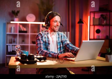 Fokussierter männlicher Manager im Headset, der die Arbeitsbesprechung durch Videoanruf auf dem Laptop leitet. Junger Mann, der redet und gesturiert, während er am Tisch in der Inneneinrichtung sitzt Stockfoto